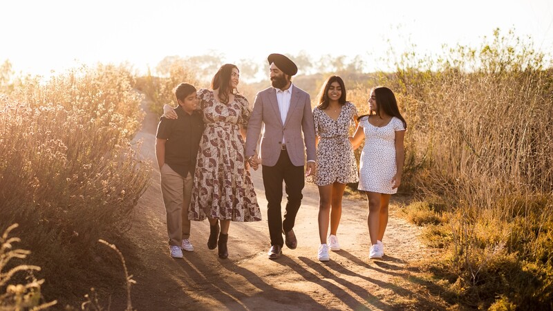 Family of five walking together on a dirt path through golden dried vegetation at sunset.