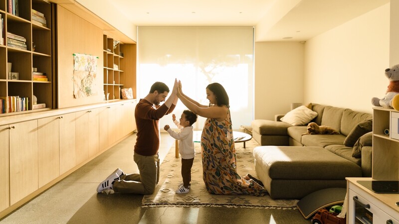 Family of three kneeling on floor with arms raised in celebration inside bright, minimalist living room.