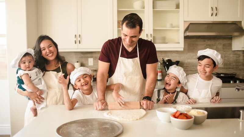 Family of six wearing aprons gathered around a kitchen counter while an adult kneads dough.