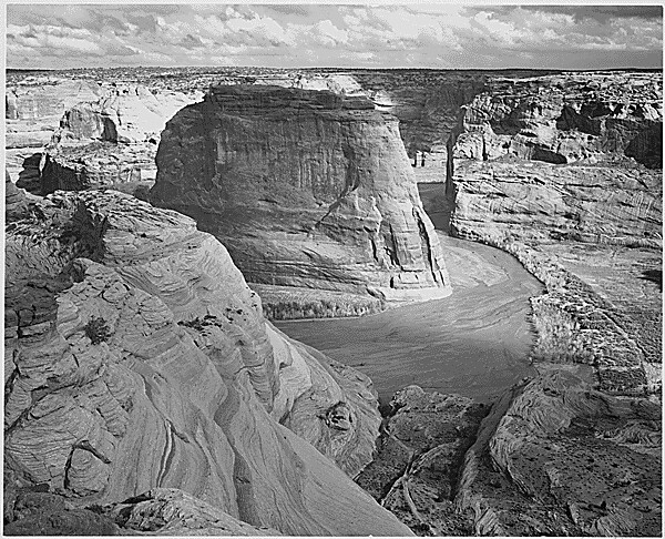 Massive butte surrounded by desert landscape with winding river in foreground under partly cloudy sky.