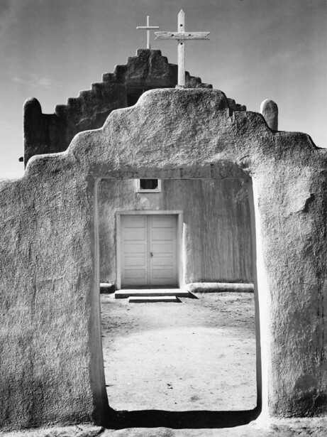 Black and white photograph of a stark adobe chapel with a cross on its roof and wooden door framing the interior.