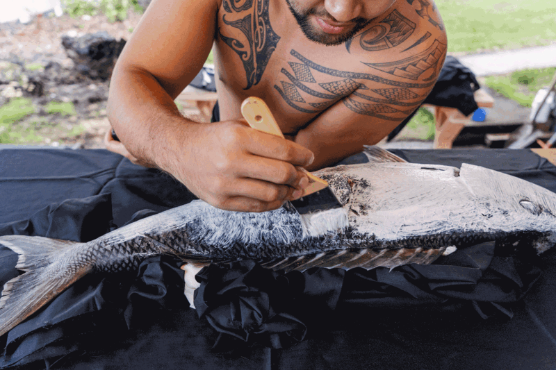Man with tribal tattoos examining a large fish on dark fabric outdoors.