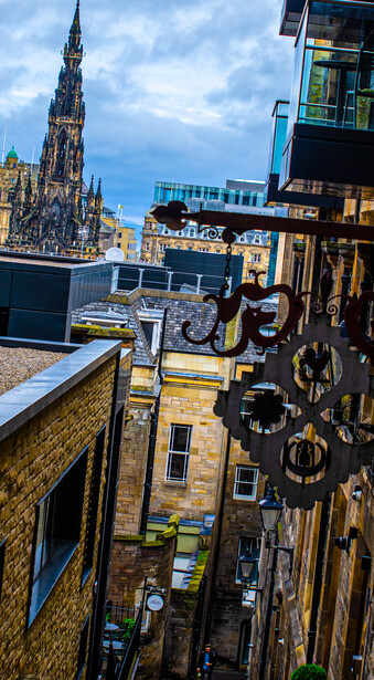 Aerial view down a narrow Edinburgh street with historic stone buildings and a Gothic spire visible in the distance.