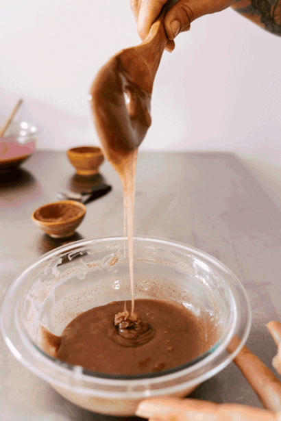 Chocolate sauce being drizzled from a wooden spoon into a glass bowl of melted chocolate.