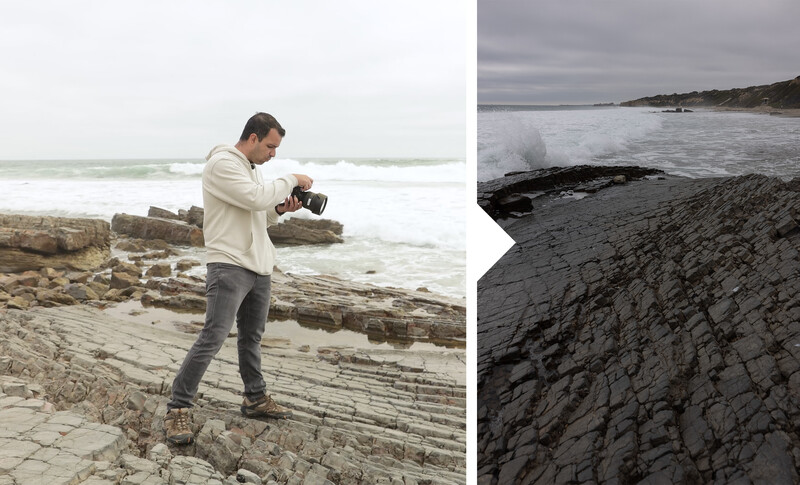 Photographer holding a camera while standing on driftwood at a rocky beach with waves in the background.