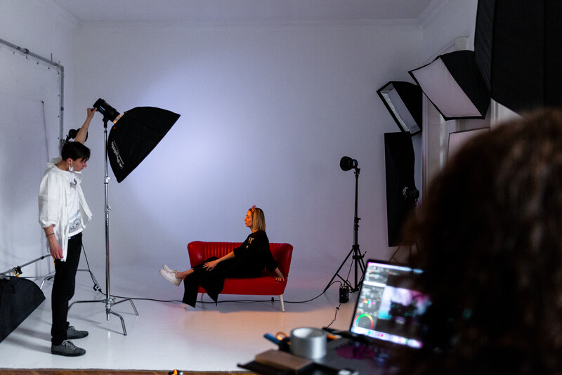 Professional studio setup with softbox lighting, subject seated on red couch, and monitor displaying editing software in foreground.