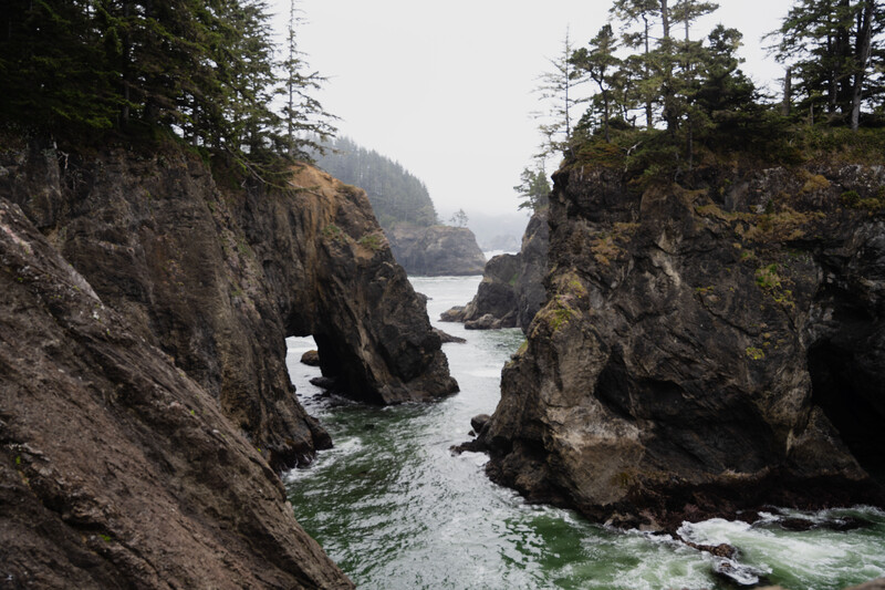 Dramatic rocky sea arch frames distant forested coastline and calm waters below.