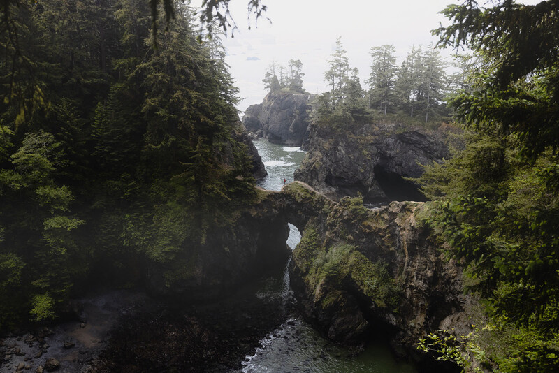 Rocky coastal landscape with moss-covered stone arches framing a turquoise river and forested headland.