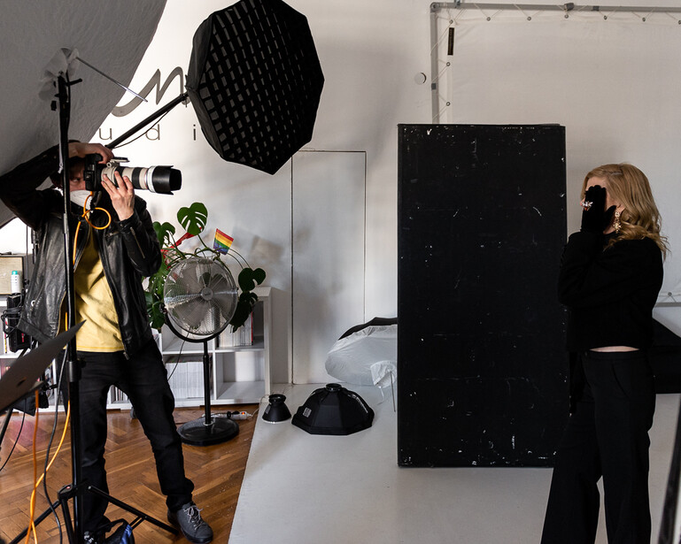 Professional studio setup with photographer adjusting camera on tripod under large softbox while assistant holds black reflector panel.