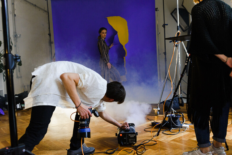 Photographer bending over camera equipment on wooden floor during studio shoot with blue backdrop and lighting rigs.