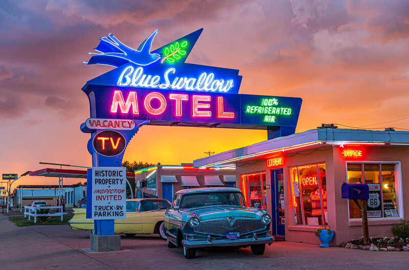 Vintage turquoise car parked in front of an illuminated retro motel sign at sunset.