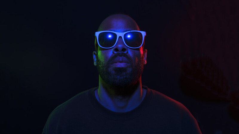 Man wearing illuminated blue-framed glasses against a dark background with red and blue gel lighting.
