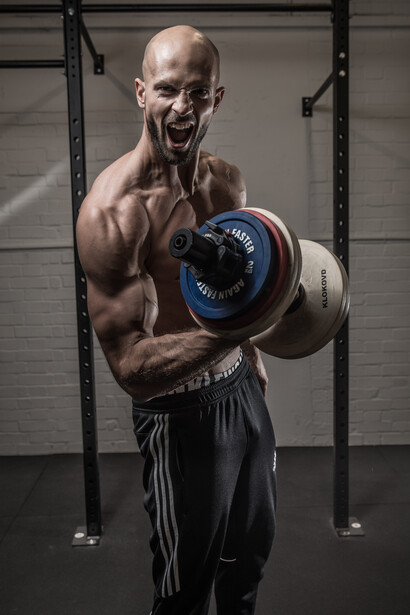 Shirtless male athlete flexing bicep while holding a dumbbell in a gym setting.