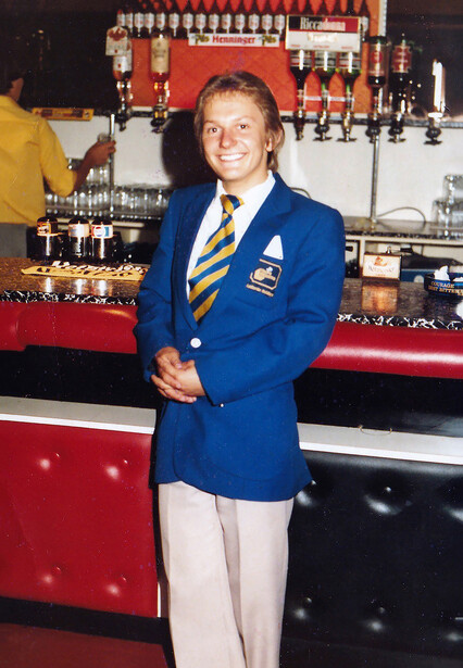 Woman in blue blazer and white pants standing in front of a bar with bottles and holiday decorations.