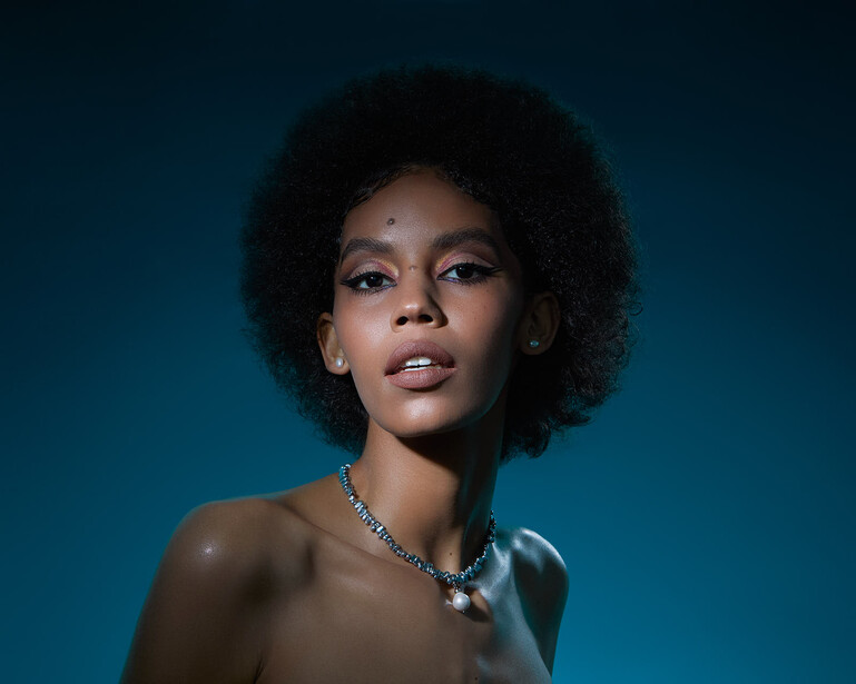 Woman with natural textured hair wearing a diamond necklace against a teal blue background.
