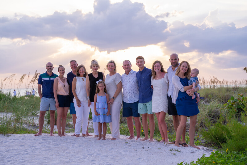 Large multi-generational family group posing together on a beach at golden hour with dramatic sky above.