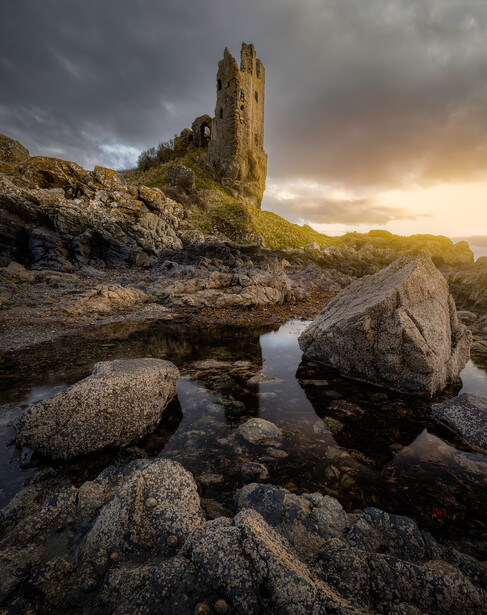 Stone tower ruins on a hillside with rocky water features in foreground at golden hour.