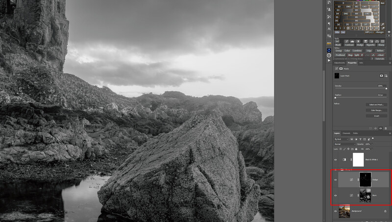 Black and white landscape showing large boulders beside still water with forested mountains in misty background.