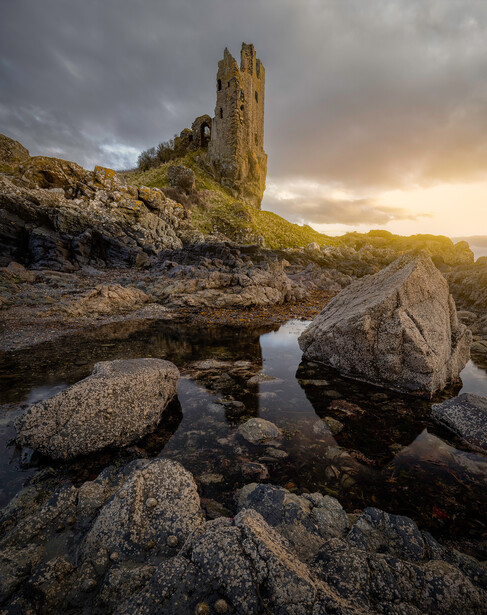 Stone tower ruins on a hillside reflected in a shallow pool of water among rocky boulders.