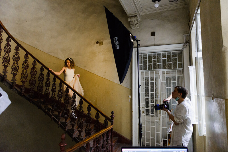 Woman on yellow stairwell landing poses while photographer with softbox lighting captures portrait in architectural interior.