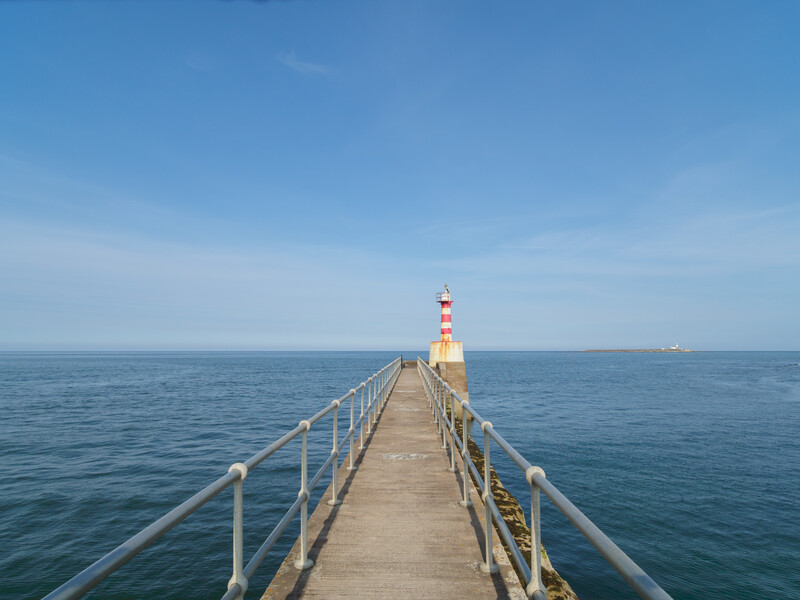 Long wooden pier extending into calm blue ocean water toward a red and white striped lighthouse under clear sky.