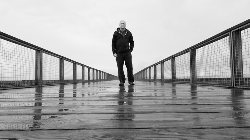 Person in black jacket standing centered on wet boardwalk flanked by metal mesh railings extending into distance.