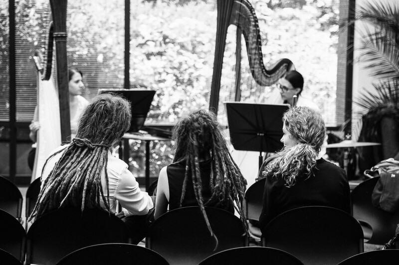 Three audience members seated from behind watching performers with harps on stage in black and white.
