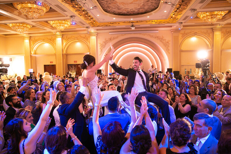 Bride and groom lifted on shoulders by wedding guests in an ornate ballroom during reception.