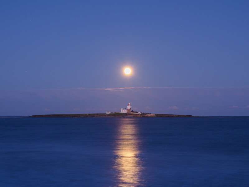 Full moon rising above a distant lighthouse on an island at twilight, with moonlight reflecting across calm water.