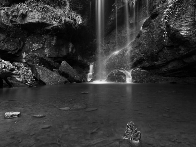 Waterfall cascading into a dark pool surrounded by moss-covered rocks in black and white.
