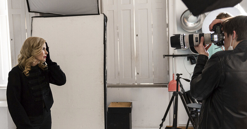 Cameraman filming a woman in black jacket against white studio backdrop with professional lighting setup.