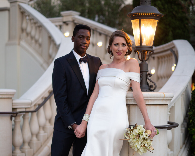 Couple in formal evening wear posing on an elegant stone balcony at dusk.