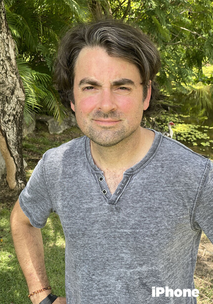 Man with dark hair wearing a gray henley shirt outdoors against green foliage.