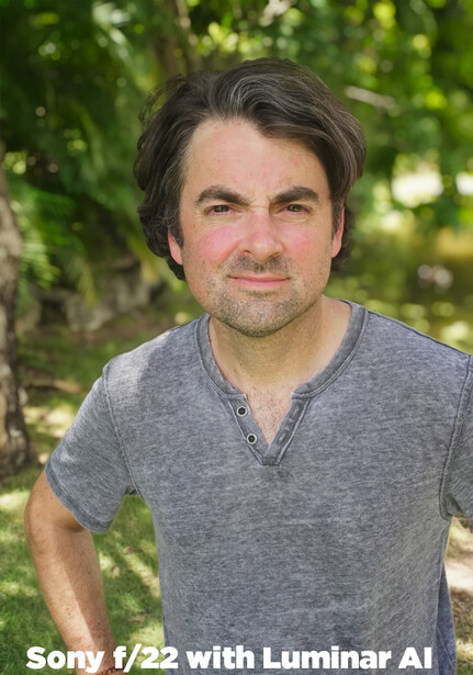 Man with dark hair wearing gray henley shirt outdoors with blurred green foliage background.