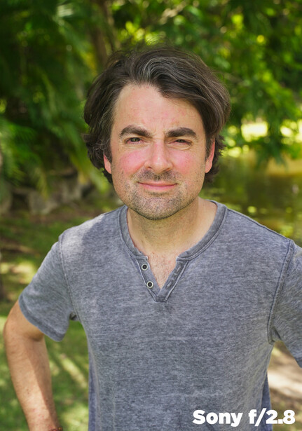 Man with shoulder-length dark hair wearing a gray henley shirt against a blurred green foliage background.