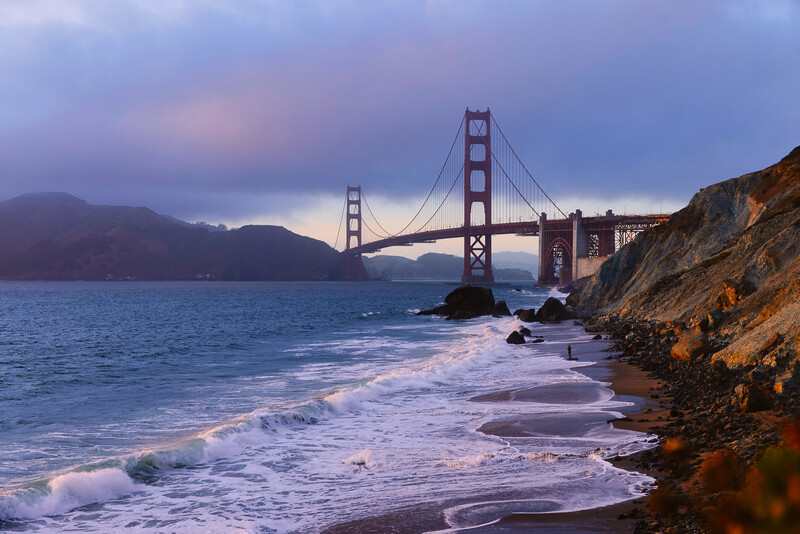 Suspension bridge spanning across a bay at twilight, photographed from a rocky beach with waves in the foreground.