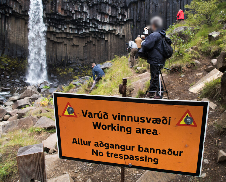 Orange warning sign in Icelandic and English blocking access to a working area near a waterfall and basalt columns.