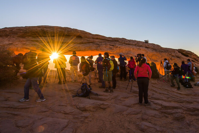Large group of photographers and spectators positioned on rocky terrain during golden hour, with intense sunburst creating lens flare.