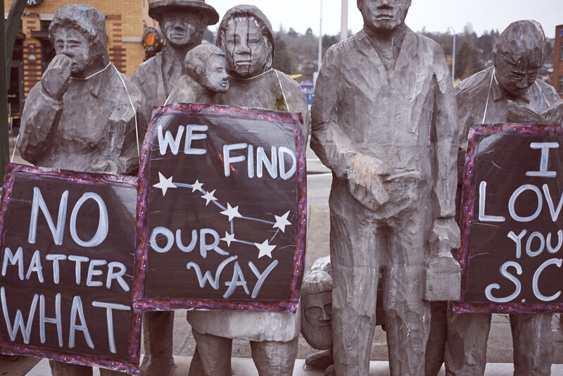 Stone statues holding protest signs with handwritten messages about unity and inclusivity.