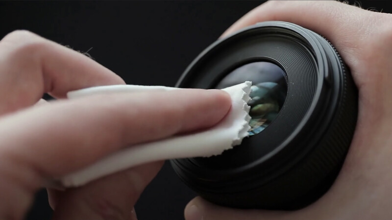 Hand cleaning a camera lens with a soft cloth against a dark background.