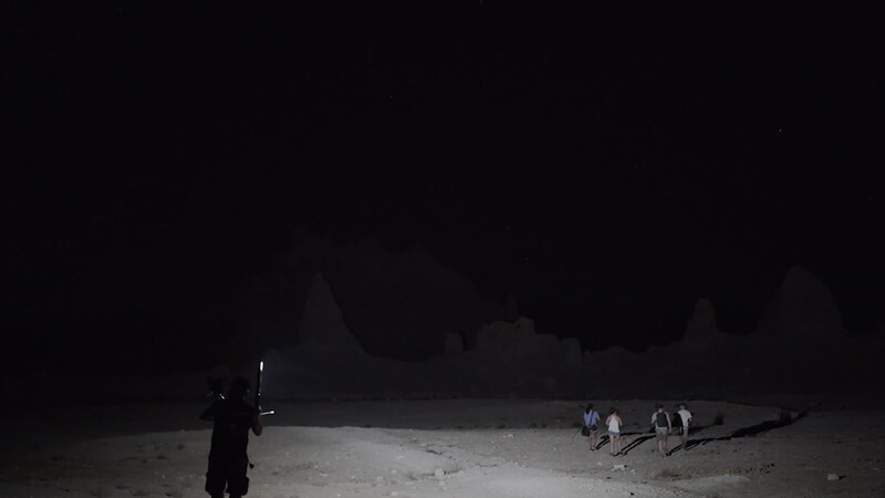 Silhouetted figure standing alone on a dark beach at night while distant people gather near the shoreline.
