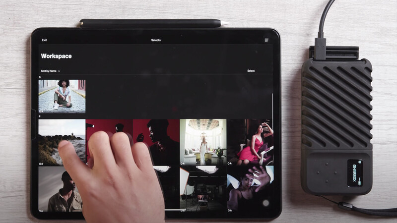 Tablet displaying a workspace with photo grid while a hand gestures at the screen, with external hard drive visible beside it.