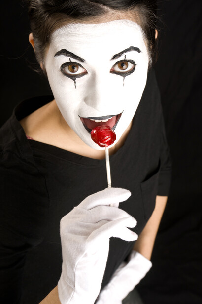 Mime performer with white face makeup holding red lollipop against black background.
