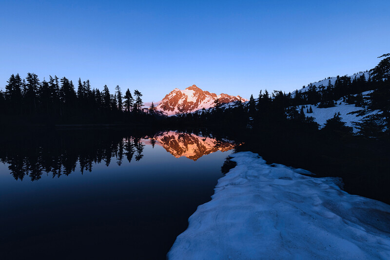 Snow-covered shoreline with perfectly still lake reflecting snow-capped mountain peak at dawn.