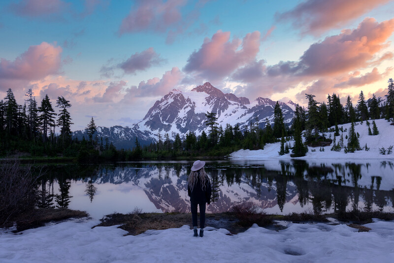 Solitary figure standing on snowy shore facing snow-capped mountain reflected in still lake at sunset.