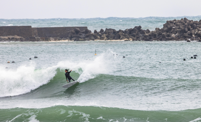 Surfer riding a green wave with rock jetty visible in the background.