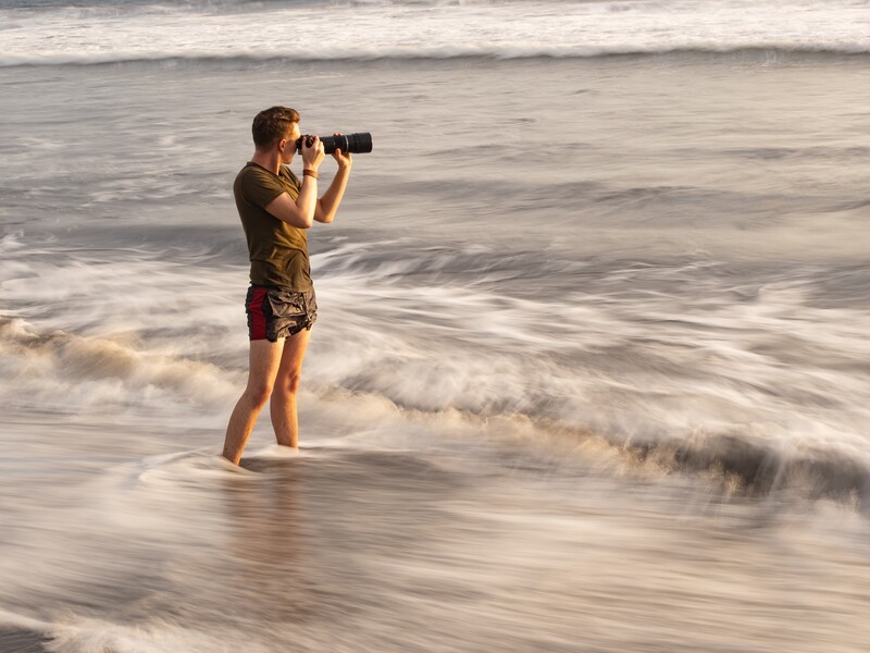 Photographer standing in shallow ocean waves while holding a telephoto lens camera to their eye.