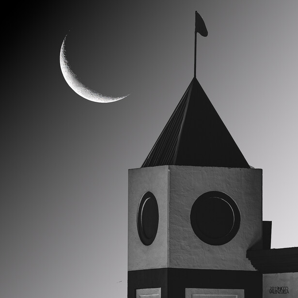 A crescent moon hangs above a modernist tower with circular windows and a flag on top, photographed in high-contrast black and white.