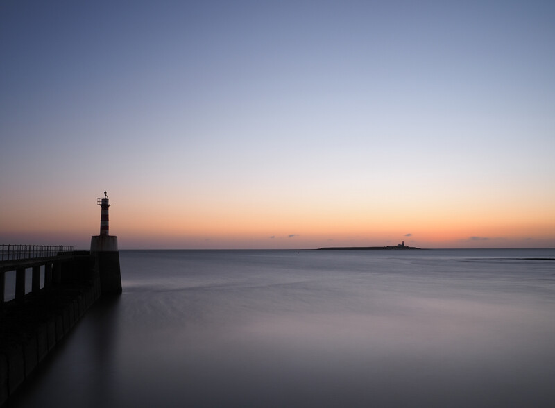 Silhouetted lighthouse and pier structure against a twilight sky with calm water and distant sailboat.