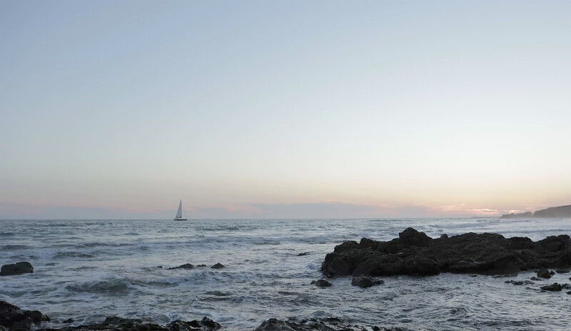 Sailboat on calm waters at dusk with rocky coastline in foreground.